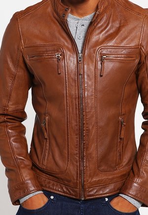 Veste en cuir marron avec fermeture éclair devant, quatre poches zippées, portée sur un t-shirt henley gris et un jean bleu foncé.