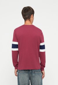 Long-sleeve, burgundy t-shirt with navy and white striped accents on sleeves. Soft fabric with a relaxed fit, featuring a round neckline.