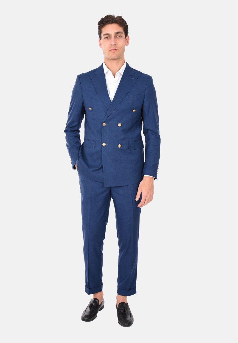 Abito blu doppiopetto con bottoni dorati, vestibilità su misura, rever a becco e pantaloni slim. Un paio di scarpe eleganti nere completa il look.