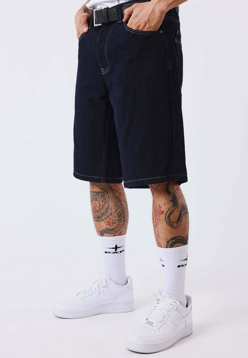 Homme portant un short en jean noir, une ceinture blanche, des chaussettes hautes blanches avec un logo noir, des baskets blanches et des tatouages visibles sur les jambes sur un fond uni.