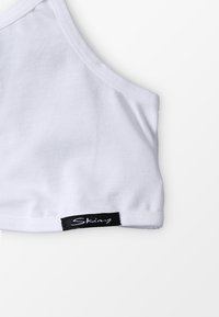 Biely bavlnený bralet s hladkým povrchom, s prešitým okrajom a čiernou nálepkou s nápisom "Skiny" v písanom štýle na spodnej strane.