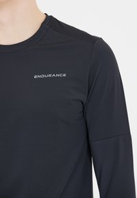 Schwarzes langärmliges Sportoberteil mit Rundhalsausschnitt. Hat eine glatte Textur und ein reflektierendes "ENDURANCE"-Logo über der Brust.