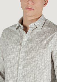 Camicia bianca con bottoni, caratterizzata da un sottile motivo geometrico ripetuto in grigio chiaro. Il tessuto è liscio e presenta un design con colletto classico.
