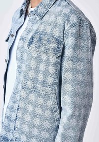 Veste en denim bleu clair avec un motif en diamant matelassé, dotée d'un col rabattu et de poches avant avec boutons. Texture lisse.