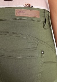Pantalones verde oliva con una textura suave, que presentan un parche de cuero etiquetado como "ONLY" y costuras decorativas en los bolsillos.