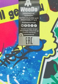Värikäs kankaan etiketti, jossa on sekoitus sinisiä, keltaisia, vaaleanpunaisia ja vihreitä graffitomaisia kuvioita. Tekstissä on hoito-ohjeet ja brändäys.