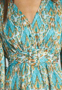 Robe avec un motif abstrait turquoise et beige, décolleté en V profond, manches longues, et une ceinture en tissu assortie avec une boucle rectangulaire.