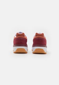 Pantofi sport roșii și portocalii cu partea superioră din suede și plasă, talpă intermediară albă și talpă exterioară din cauciuc bej, având brandingul Reebok pe călcâi.