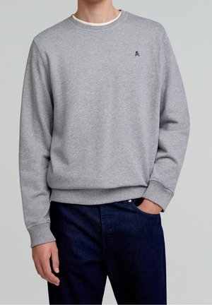 Man wearing light grey crewneck sweatshirt with small black skull logo on chest and dark blue jeans, hand in pocket.