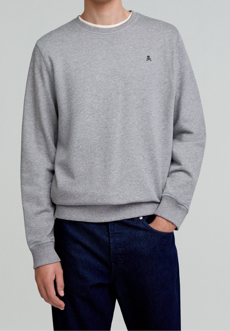 Homme portant un sweat-shirt gris clair à col rond avec un petit logo de crâne noir sur la poitrine et un jean bleu foncé, la main dans la poche.