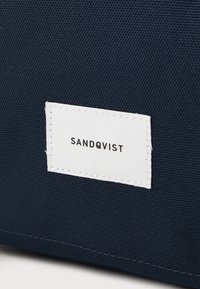 Granatowa torba z tkaniny o fakturowanej powierzchni, z białą metką wszytą z napisem "SANDQVIST" w czarnym kolorze. Prosty design, bez dodatkowych akcentów.