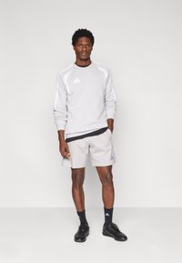 Φόρμα ανοιχτού γκρι χρώματος με λευκές λεπτομέρειες και μαύρο περίγραμμα, συνδυασμένη με ανοιχτό γκρι σορτς που έχουν μαύρη οριζόντια λωρίδα και λογότυπο. Μαύρα αθλητικά παπούτσια.