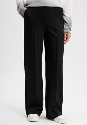 Person trägt schwarze, weit geschnittene Hose, weiße Sneaker und ein grau-weiß verlaufendes Langarmshirt, eine Hand in der Tasche.