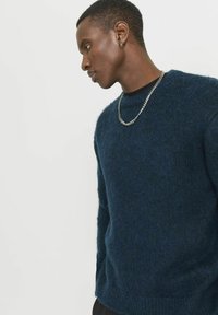 Gestrickter Pullover in dunkelmarineblau, mit rundem Ausschnitt, strukturiertem Stoff und langen Ärmeln. Kombiniert mit einer silbernen Kettenhalskette.