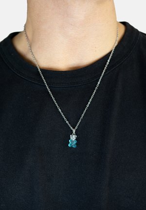 Collier en chaîne argentée avec un pendentif en forme de ours en gélatine bleu transparent, avec des détails texturés, porté sur une chemise noire.