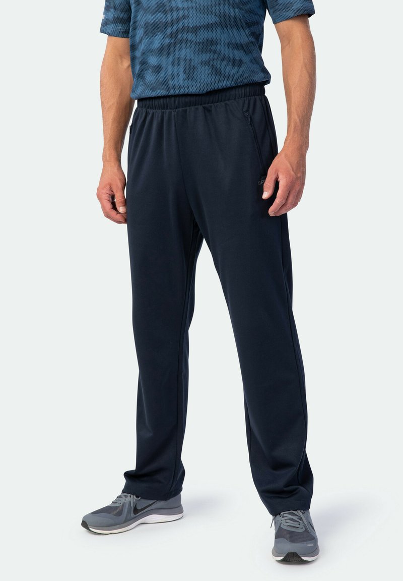 Mann in schwarzen Sporthosen mit elastischem Bund und blauem gemustertem Shirt, stehend, mit grauen Sportschuhen vor weißem Hintergrund.