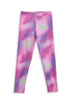 Młoda dziewczyna ubrana w różowo-fioletowy sportowy strój tie-dye z krótkim topem, kurtką na zamek, legginsami i lawendowymi trampkami, stojąca na szarym tle.
