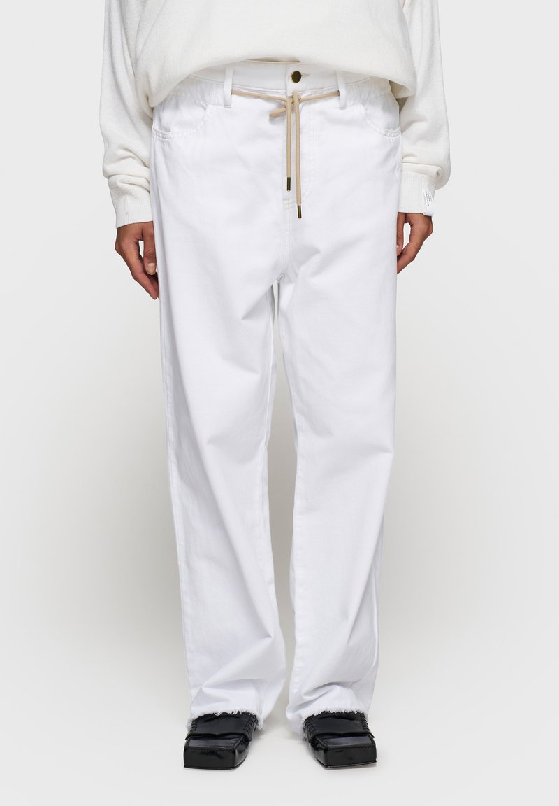 Białe bawełniane spodnie o luźnym kroju, z elastycznym pasem na sznurek, przednimi kieszeniami i postrzępionym dołem. Noszone z czarnymi butami z podszewką.