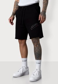 Shorts en coton noir avec une taille élastique et un cordon de serrage. Présente un texte coloré brodé sur la cuisse gauche. Associés à des baskets blanches.