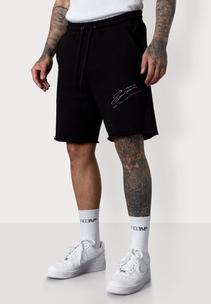 Shorts en coton noir avec une taille élastique et un cordon de serrage. Présente un texte coloré brodé sur la cuisse gauche. Associés à des baskets blanches.
