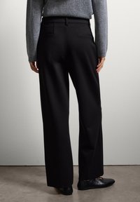Zwarte wijd uitlopende broek met een gladde textuur, twee achterzakken en een rechte snit. Gecombineerd met zwarte, laag uitgesneden schoenen.