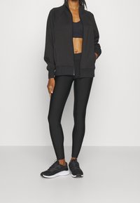 Svart zip-up jacka med raglanärmar, tillsammans med en svart sport-bh och högmidjade leggings. Svarta sneakers med texturerad detaljering.