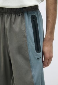Šedé a modré nohavice na jogging s bočnou zipsovou vreckom, elastickým pásom a logom Nike pri leme. Hladká textúra, uvoľnený fit a športový dizajn.