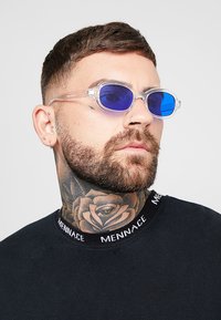 Gennemsigtige solbriller med blå linser, oval form og letvægtsdesign. Båret med en sort skjorte med grafisk kravedetalje.