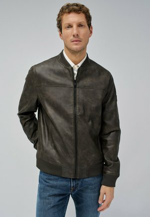 Hombre vestido con una chaqueta bomber de cuero marrón oscuro sobre una camisa blanca y jeans azules, de pie con una mano en el bolsillo contra un fondo simple.