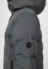 Veste grise rembourrée avec capuche. Présente une surface texturée, des poignets élastiques et un logo hexagonal noir sur la manche.