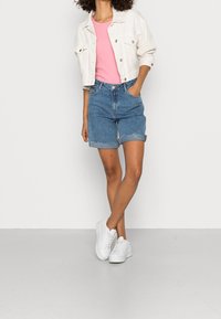 Biała dżinsowa kurtka na różowej koszulce, zestawiona z niebieskimi szortami dżinsowymi z podwiniętymi nogawkami i białymi sneakersami. Ustawiona na jednolitym tle.