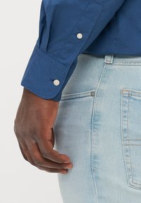 Modrá košeľa na gombíky s prispôsobeným manžetou a dvoma bielymi gombíkmi, nosená s svetlomodrými džínsy. Hladká textúra látky, bez vzorov.