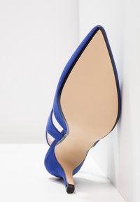 Chaussure à talon haut en daim bleu à bout pointu reposant sur le côté contre un mur d'angle blanc.