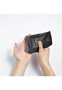 Pochette noire étiquetée "ROSEMARY COLD BREW" avec du texte blanc, en cours de compression pour déverser un produit gris sur une main.