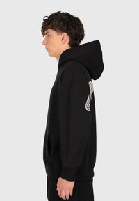 Junger Mann in schwarzem Kapuzenpullover mit hochgezogener Kapuze, der im Profil vor einem schlichten hellen Hintergrund steht.