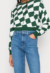 Grön och vit stickad tröja med ett geometriskt diamantmönster, kombinerad med högmidjade blå jeans. Tröjan har ribbade fållar och muddar.