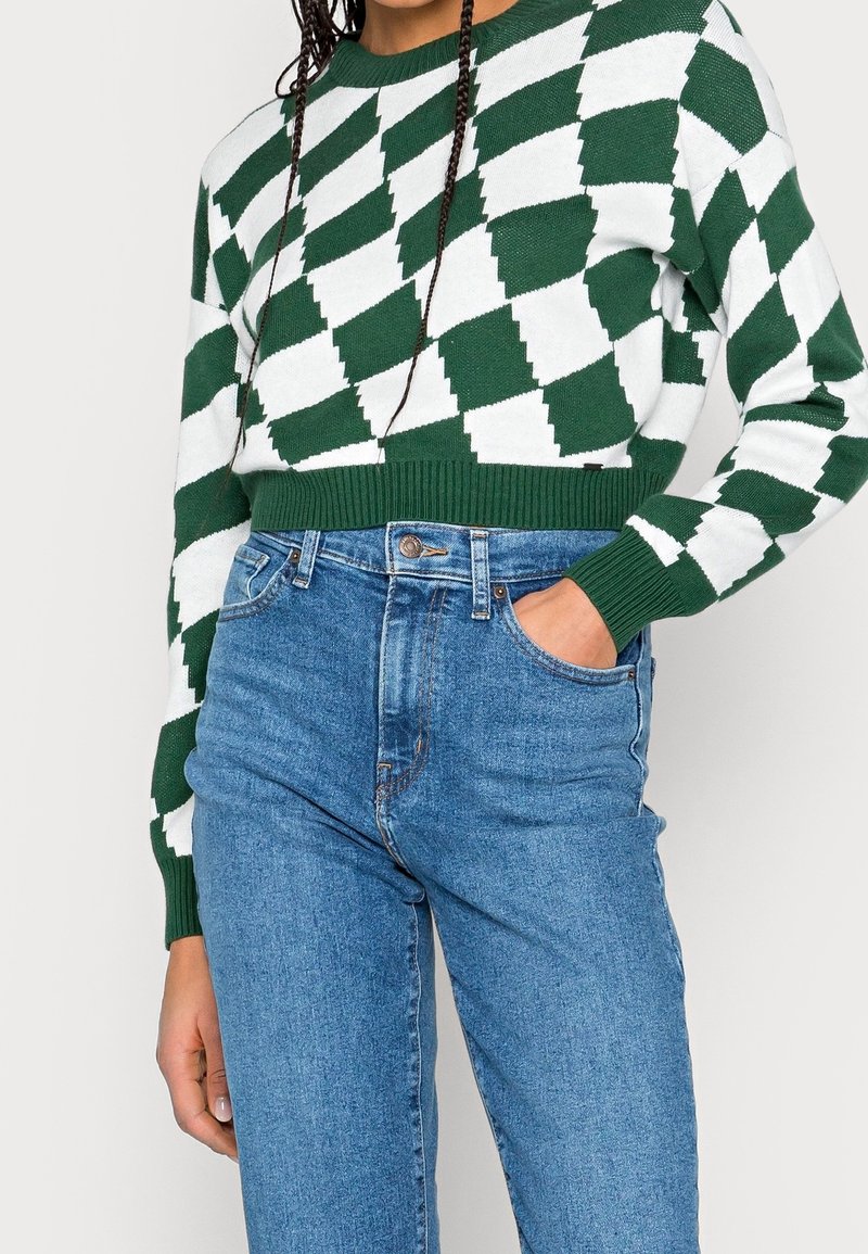 Grön och vit stickad tröja med ett geometriskt diamantmönster, kombinerad med högmidjade blå jeans. Tröjan har ribbade fållar och muddar.