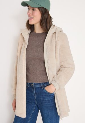 Beige fleece-gevoerde jas met een tan buitenkant, een ritssluiting aan de voorkant en een capuchon; gedragen over een bruine trui en blauwe spijkerbroek, met elastische manchetten.