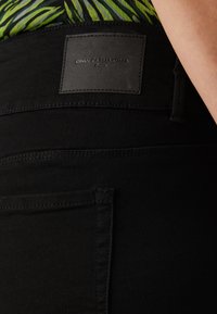 Black high-waisted jeans with a textured leather label. Features stitching details and back pockets, presented against a patterned top.