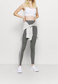 Leggings grigie con strisce laterali bianche, abbinate a un top corto bianco e a una felpa grigia chiara legata in vita. Sneakers bianche.