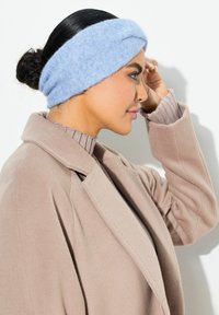Fascia per la testa di colore azzurro chiaro, lavorata a maglia e con design intrecciato, realizzata in materiale morbido, indossata con un cappotto di lana beige e un dolcevita a costine.