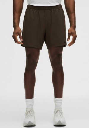 Mann trägt dunkelbraune Sportshorts, weiße Crew-Socken und weiße Laufschuhe und steht vor einem einfarbigen Hintergrund.