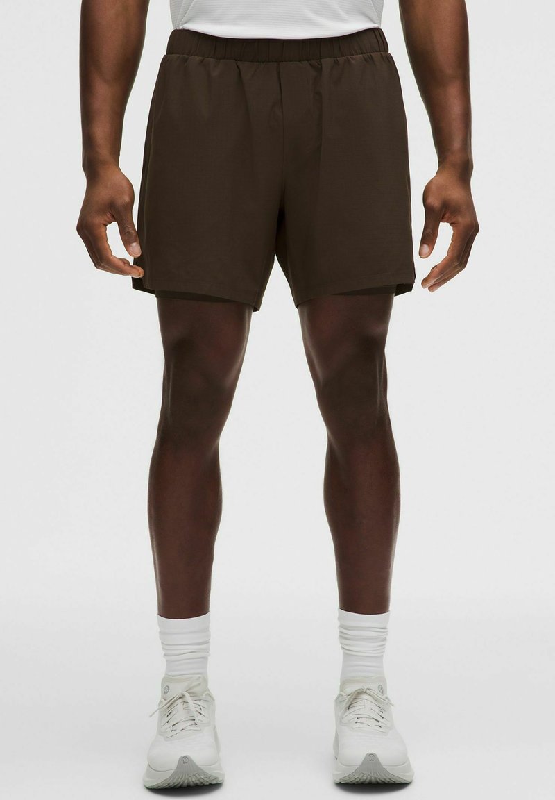 Bărbat purtând pantaloni scurți atletici maro închis, șosete albe de tip crew și pantofi albi de alergare, stând pe un fundal simplu.