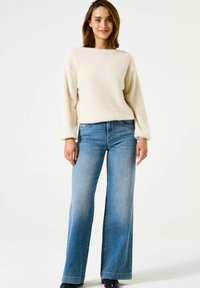 Un pull créme, fluffy avec des manches longues associé à un jean ample en délavé clair. La tenue présente des coutures minimales et des accents de denim classiques.