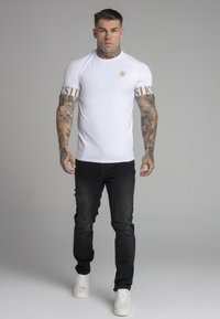 White fitted t-shirt with logo on chest and patterned sleeves; paired with black jeans and white sneakers, featuring a distressed knee detail.
