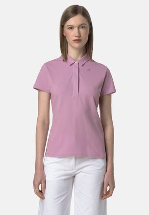 Donna che indossa una polo rosa a maniche corte e pantaloni bianchi, in piedi davanti a uno sfondo chiaro e uniforme, rivolta in avanti.