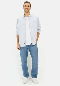 Jasnoniebieska koszula z guzikami nałożona na białą koszulkę, w połączeniu z niebieskimi dżinsami i białymi trampkami, z kieszenią z przodu i luźniejszym krojem.