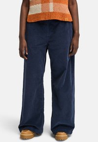 Pantalones de pana anchos en color azul marino con una textura suave, que presentan un corte relajado y una ligera estría vertical, acompañados de calzado en color marrón claro.