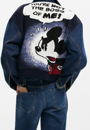 Veste en denim cropped avec un motif d'un personnage de dessin animé au dos, accompagné de texte en gras au-dessus. Teintes bleu foncé et bleu clair, poignets côtelés.