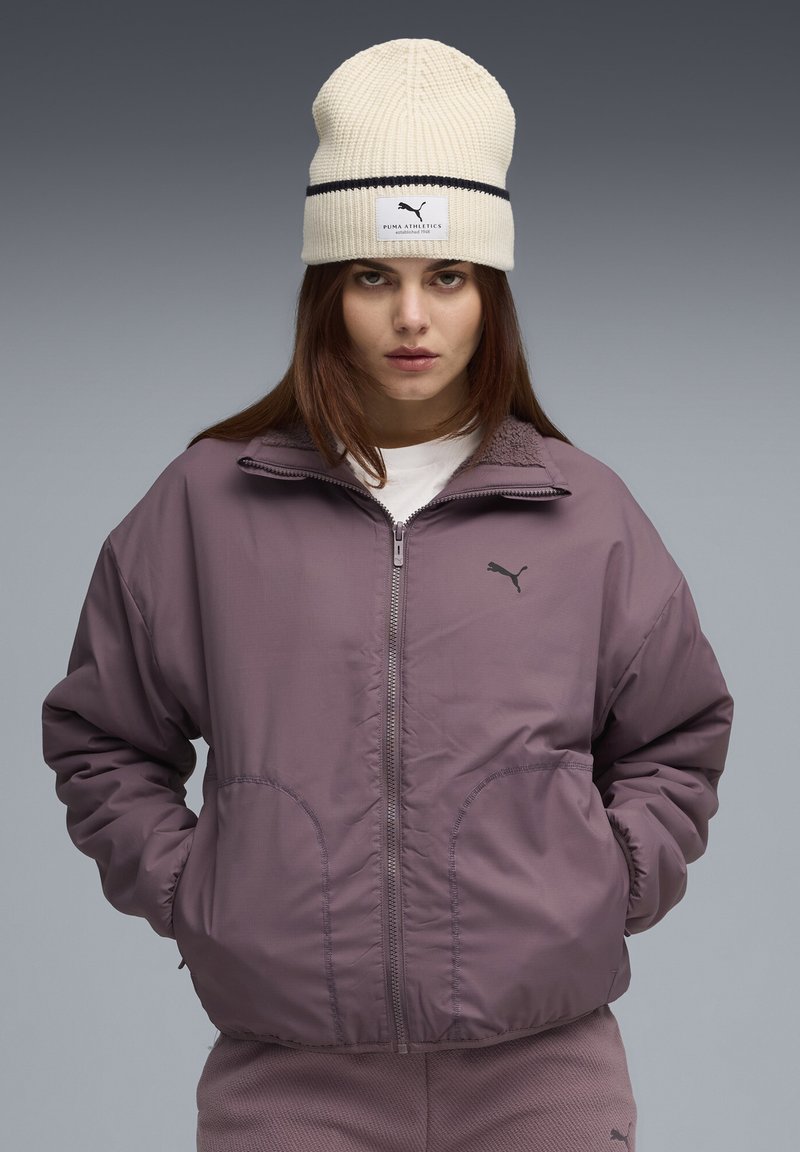 Giacca imbottita viola con tasche laterali e cerniera, abbinata a un berretto di maglia crema con una banda nera e un'etichetta con logo.